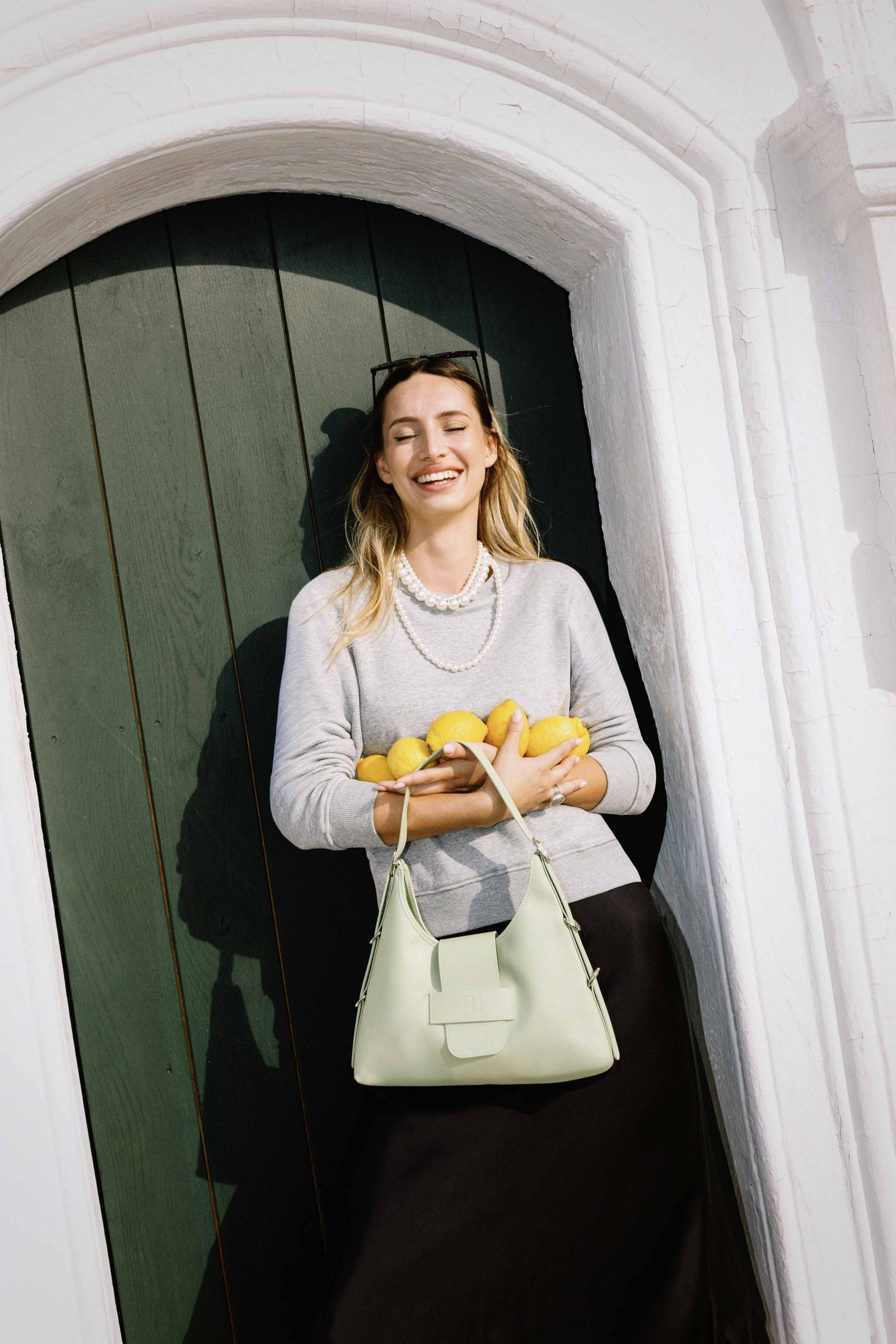 une jeune femme devant une porte fermée porte à la main le sac fada taille M vert clair. elle a aussi dans sa main des citrons